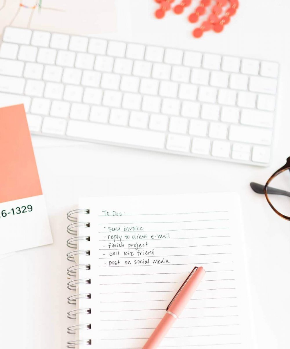 Desk setup with a keyboard, pink pen, glasses, and notepad listing tasks. Coral design swatches add a pop of color.